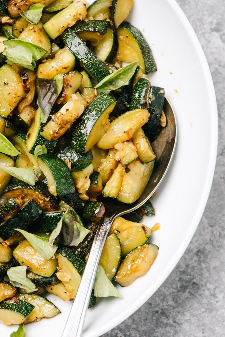 A serving spoon tucked into a white serving bowl of crisp sautéed zucchini.