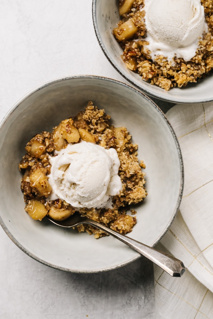 Two bowls of healthy apple crisp topped with vanilla ice cream on a cement background with a white and gold linen napkin.