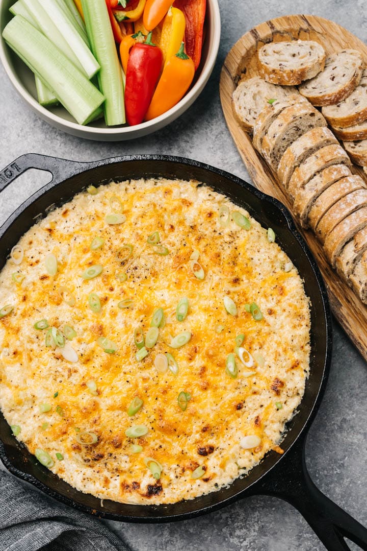 Hot crab dip in a cast iron skillet surrounded by sliced bread and vegetables on platters.