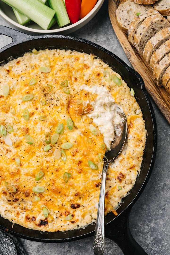 Hot maryland crab dip in a cast iron skillet with silver serving spoon surrounded by sliced vegetables and bread slices.
