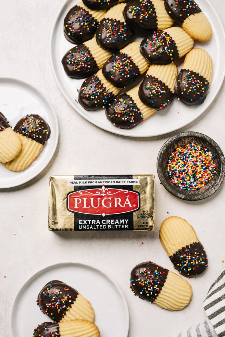 A block of Plugra butter surrounded by small plates and a platter of italian butter cookies with a dish of sprinkles and a striped linen napkin.