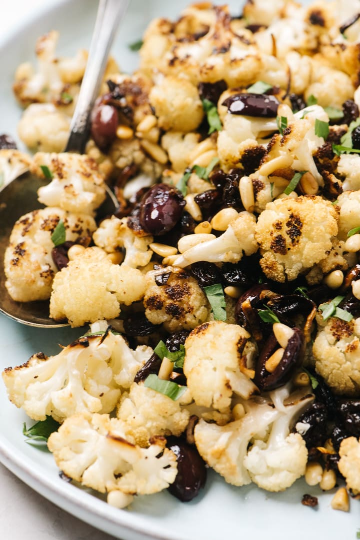 Side view, a platter of mediterranean style roasted cauliflower with olives, raisins, and pine nuts.