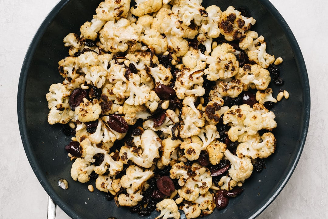 Roasted cauliflower in a skillet tossed with olives, raisins, and pine nuts.
