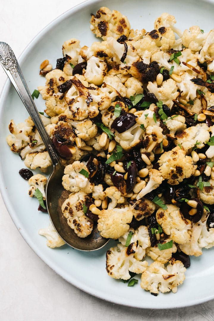 A blue serving platter of mediterranean roasted cauliflower with a silver serving spoon.
