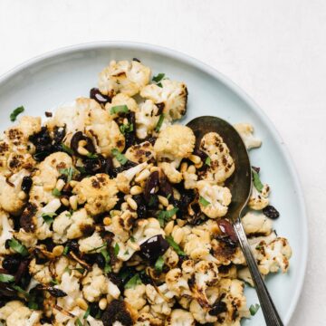 Mediterranean style roasted cauliflower with olives, raisins, and pine nuts on a blue serving platter with a silver spoon.