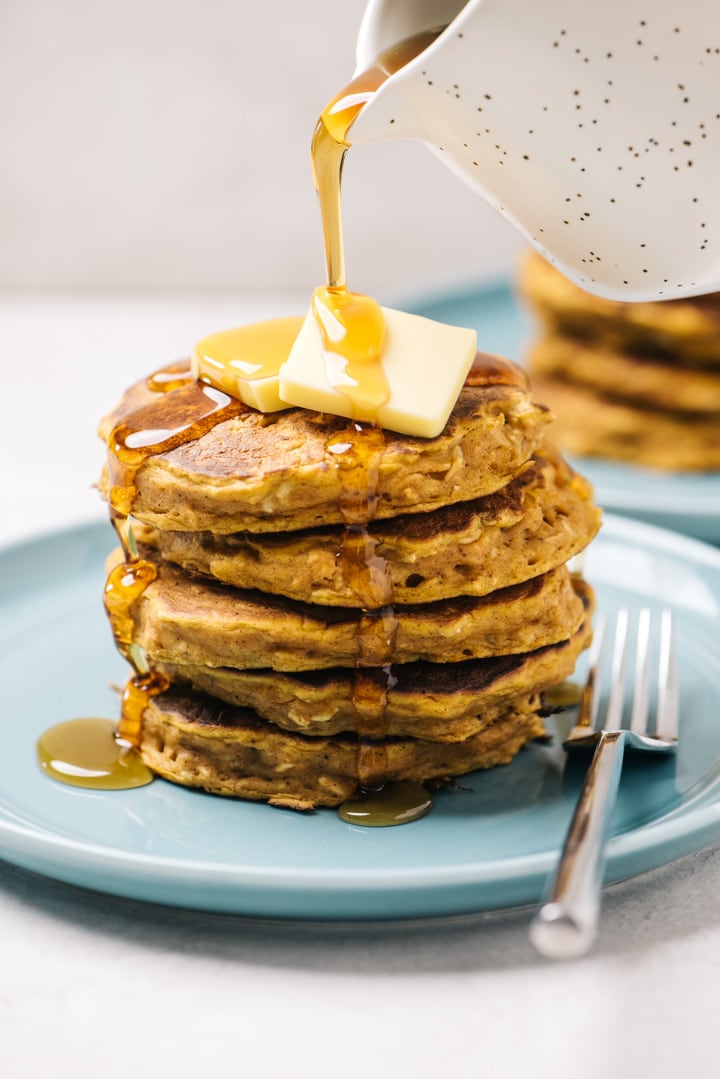 Pouring maple syrup over a stack of pumpkin oatmeal pancakes on a blue plate.