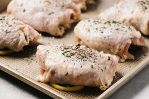 Side view, chicken thighs seasoned with rosemary positioned over lemon slices on a baking sheet.