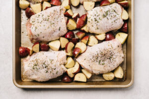 Chicken thighs seasoned with fresh rosemary arranged with potatoes on a baking sheet.