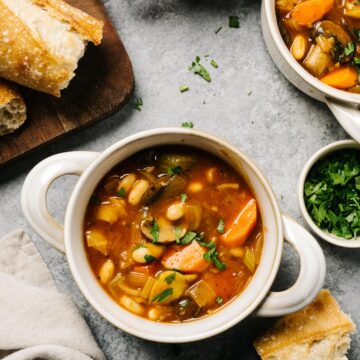 Several bowls of vegan stew on a cement background with slices of bread on a cutting board to the side and a tan linen napkin.