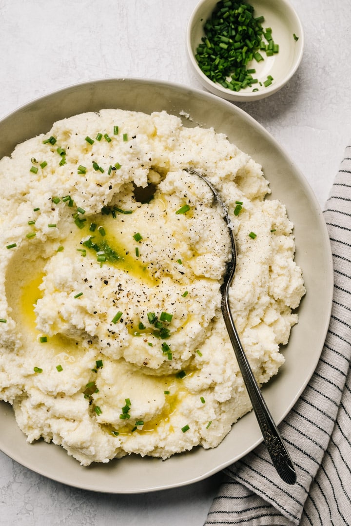 A bowl of cauliflower mashed potatoes garnished with melted butter and chives on a cement background with a striped linen napkin.