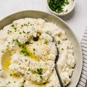 A bowl of cauliflower mashed potatoes garnished with melted butter and chives on a cement background with a striped linen napkin.