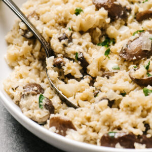 Side view, cauliflower risotto with sauteed mushrooms in a white serving bowl with a vintage silver serving spoon.