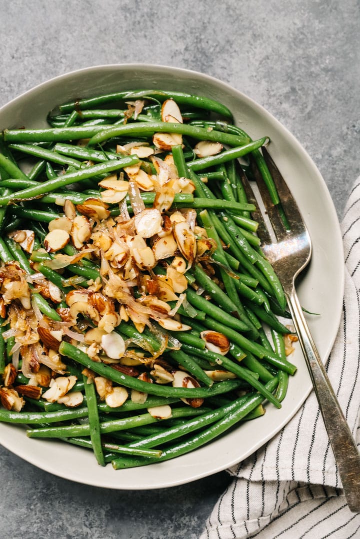 A serving bowl of green beans almondine with a silver serving fork and striped linen napkin.