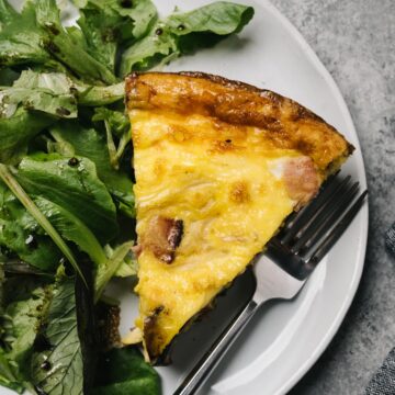 A slice of ham frittata on a white plate with a tossed green salad and silver fork.