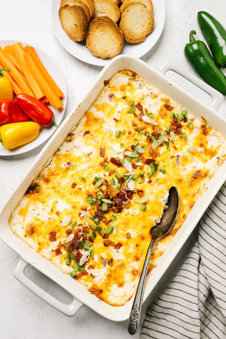 Jalapeno popper dip in a casserole dish on a concrete background surrounded by plates of bread slices and raw vegetables.