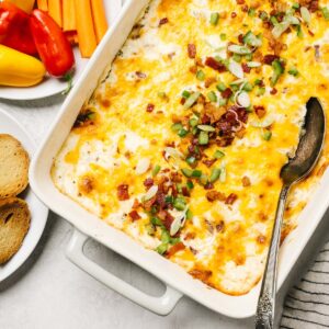 Jalapeno popper dip in a casserole dish on a concrete background surrounded by plates of bread slices and raw vegetables.