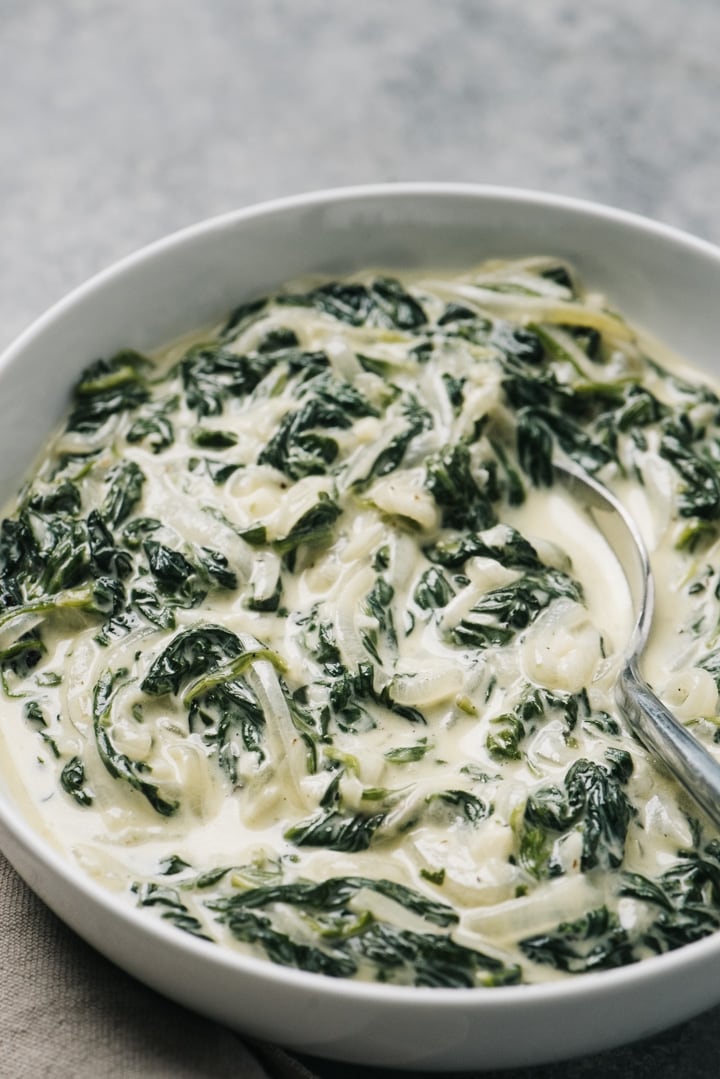 A bowl of keto creamed spinach on a cement background with a silver serving spoon and a tan linen napkin.