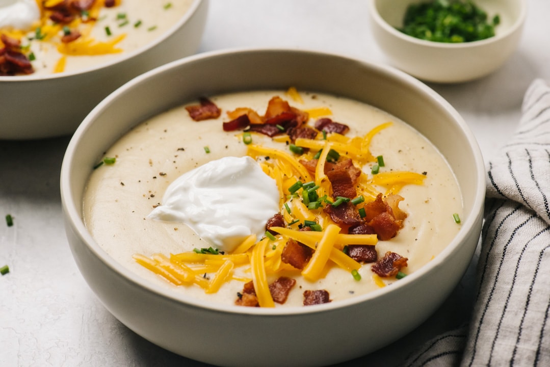 Side view, two bowls of baked potato soup topped with bacon, shredded cheese, sour cream, and chives with a linen napkin and small bowl of chives in the background.