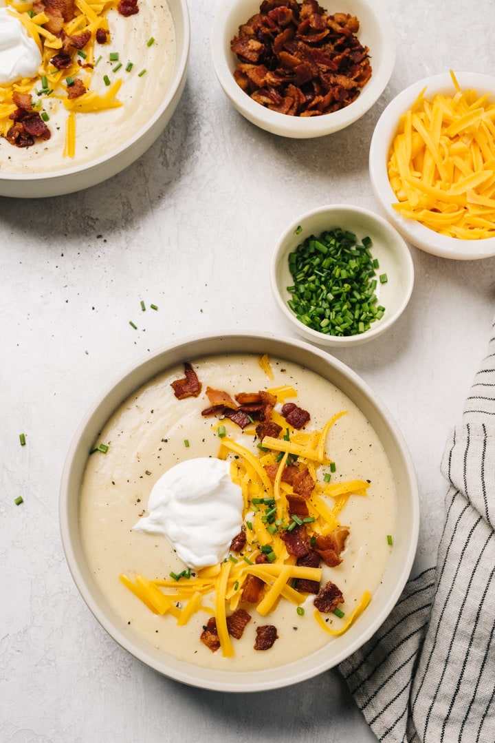 Two bowls of loaded baked potato soup topped with bacon, shredded cheese, sour cream, and chives on a concrete background with a striped linen napkin.