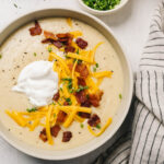 A bowl of baked potato soup topped with bacon, shredded cheese, sour cream, and chives on a concrete background with a striped linen napkin.