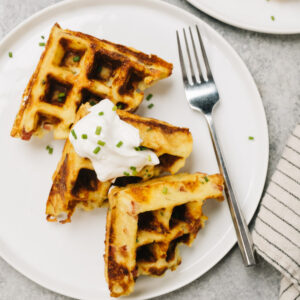 Three mashed potato waffles lined up on a white plate with a dollop of sour cream, silver fork, and striped linen to the side.