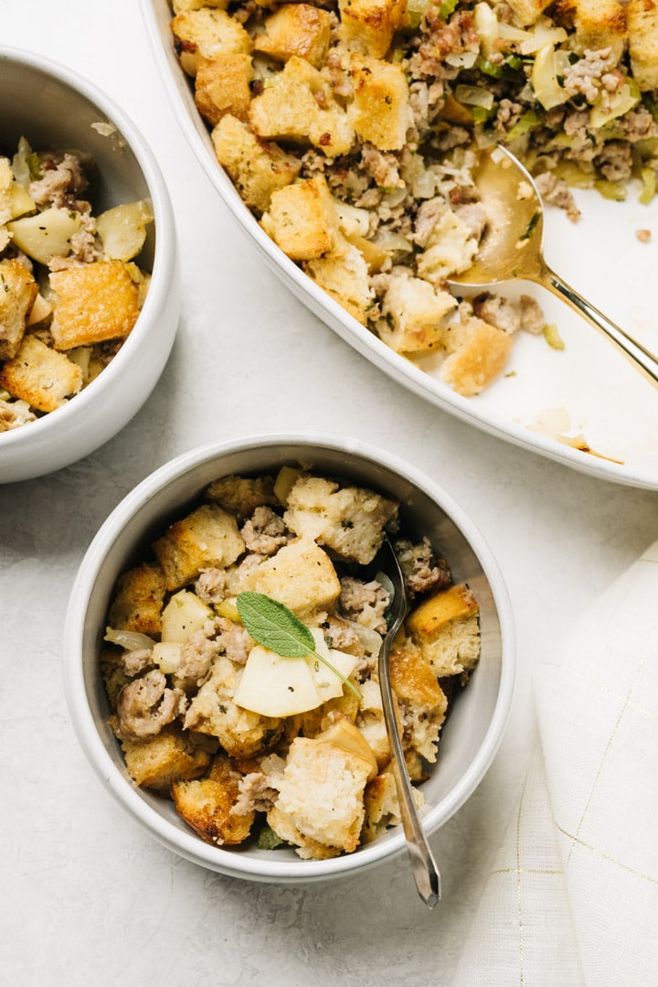 Two bowls of sausage stuffing on a concrete background with a casserole dish and linen napkin to the side.