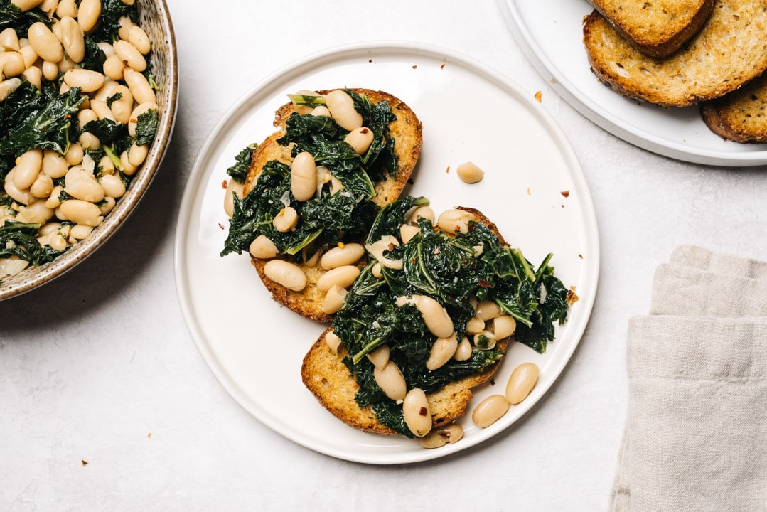 Two pieces of garlic toast topped with beans and greens on a white plate on a cement background wit a linen napkin and serving bowl to the side.