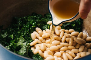 Pouring broth into a pot with wilted kale and white beans.