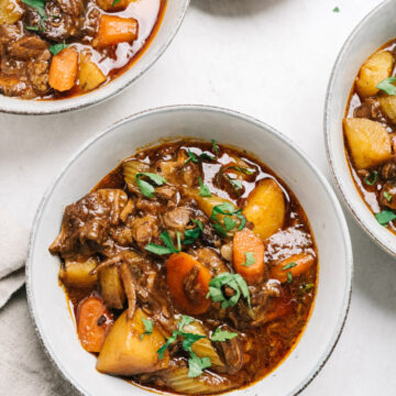 Three bowls of beef stew on a cement background with a tan linen napkin and small garnish bowl of chopped parsley.