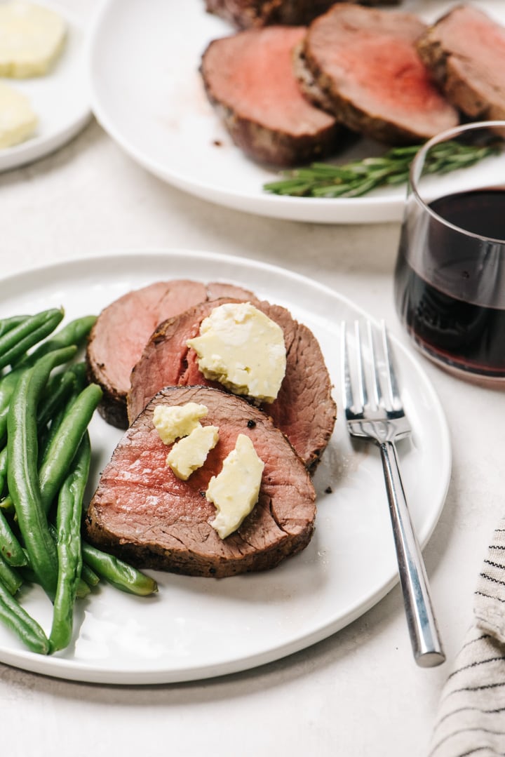 Slices of beef tenderloin topped with blue cheese butter and served with green beans on white plates with a glass of red wine and striped linen napkin.