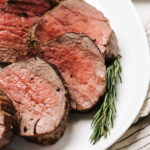 Slices of beef tenderloin sliced into ½" pieces and arranged on a white platter with a sprig of rosemary.