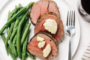 Three slices of beef tenderloin topped with blue cheese butter and served with green beans on a white plate with a silver knife and fork, glass of red wine, and striped linen napkin.