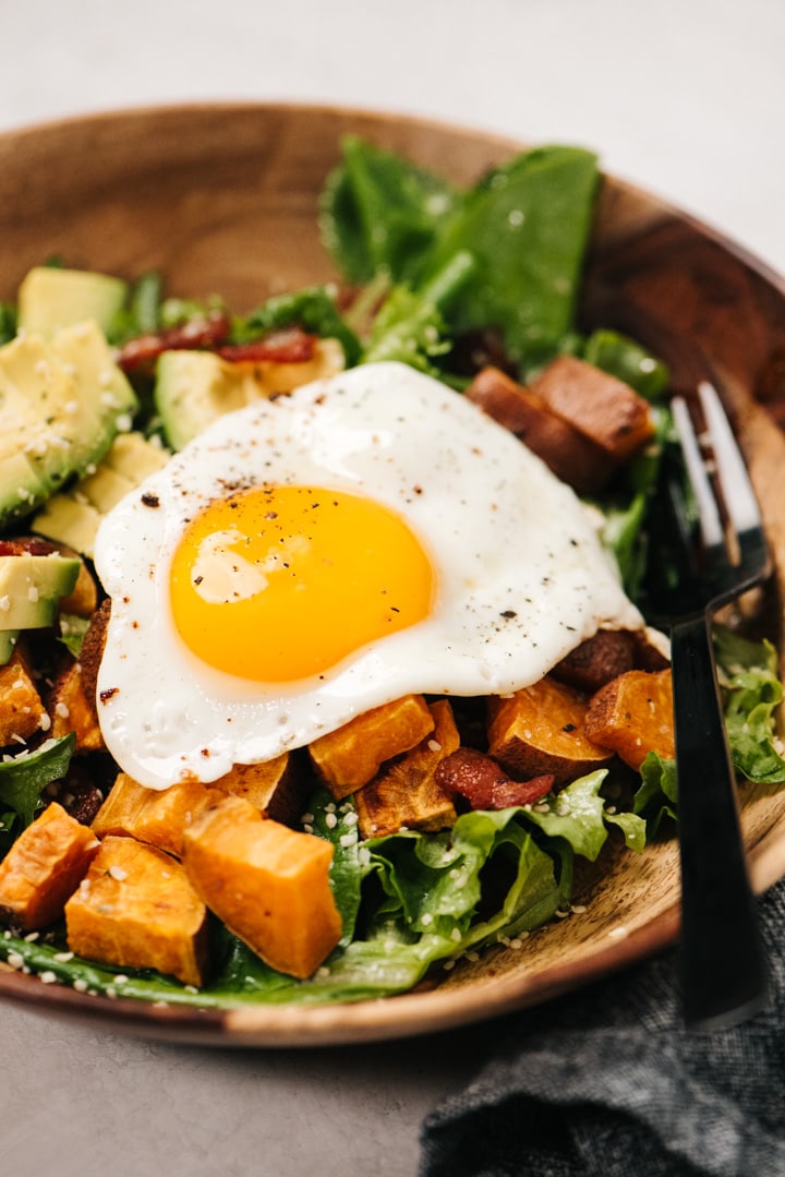 Side view, breakfast salad in a wood bowl with bacon, roasted sweet potatoes, avocado, and a fried egg.