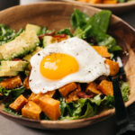 Side view, breakfast salad in a wood bowl with bacon, avocado, sweet potato, and a fried egg in a wood bowl.