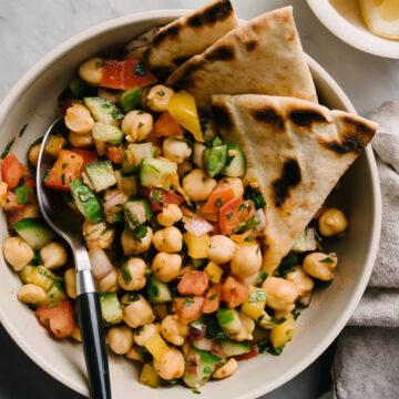 A bowl of mediterranean chickpea salad with pita bread and a spoon tucked into the bowl on a marble table.