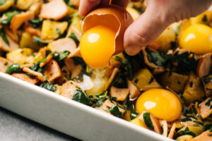 A woman's hand pouring a whole egg into a depression in a paleo eggs benedict casserole.