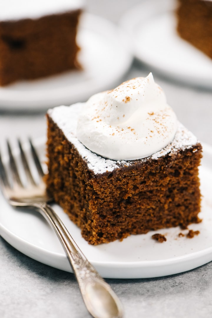 A slice of gingerbread cake topped with whipped cream and powdered sugar with a silver vintage fork.