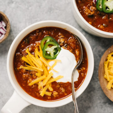 A bowl of instant pot chili topped with shredded cheese, sour cream, and sliced jalapeno on a concrete background.