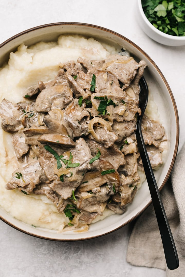 Keto beef stroganoff served over cauliflower puree in a black and cream bowl with a black fork and tan linen napkin.