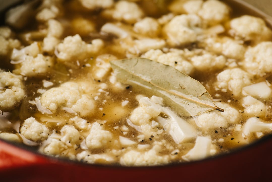 Cauliflower florets simmering with onion, leek, and broth and a bay leaf on top.