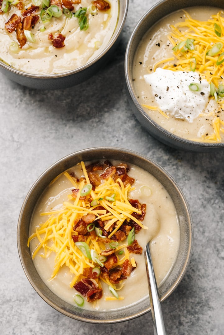 Three bowls of whole30 and keto cauliflower soup garnished with various toppings, including cheese, bacon, and sour cream, on a cement background.