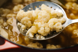 Cooked cauliflower on a slotted spoon hovering over a pot of unpureed cauliflower soup.