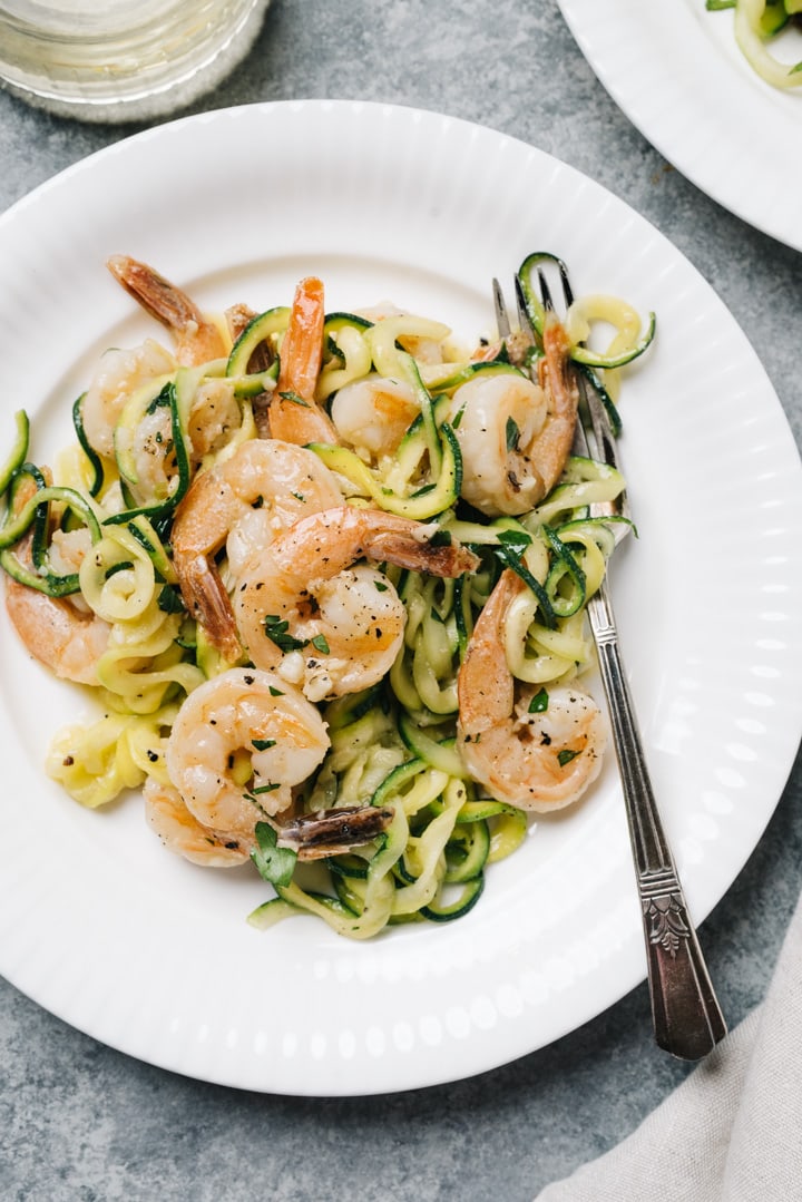 A plate of keto shrimp scampi tossed with zoodles on a white plate with a vintage fork, a wine glass to the side, and a cream colored linen napkin.