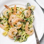 A plate of keto shrimp scampi tossed with zoodles on a white plate with a vintage fork, a wine glass to the side, and a cream colored linen napkin.
