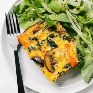 A slice of spicy keto breakfast casserole on a white plate with mixed greens and black fork.