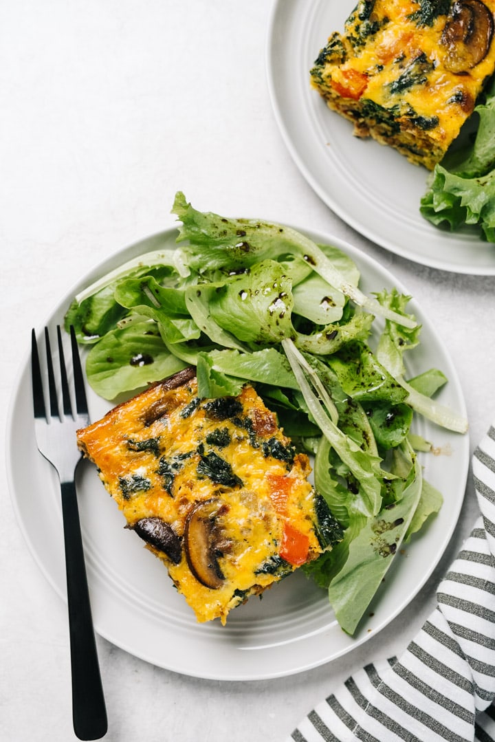 Two servings of keto breakfast casserole on a white plate, served with mixed greens, on a cement background.