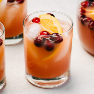 Side view, three glasses of whiskey punch next to a glass pitcher on a cement background; each cocktail is garnished with fresh cranberries and orange slices.