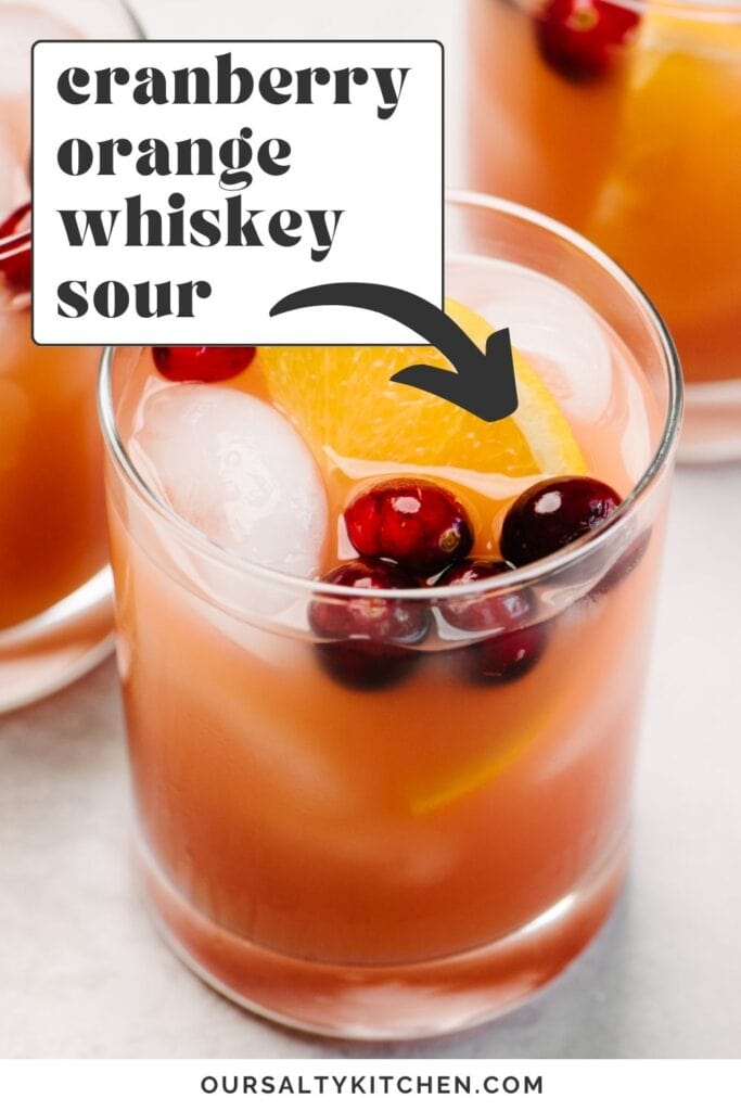 Side view, a glass of whiskey punch made with cranberry and orange juice; another cocktail and a glass pitcher are in the background; text overlay reads "cranberry orange whiskey sour".
