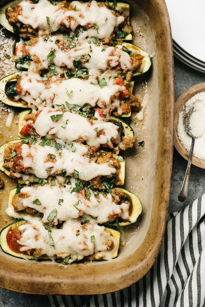 A row of stuffed zucchini in a casserole dish with a side of parmesan cheese and a striped linen napkin.