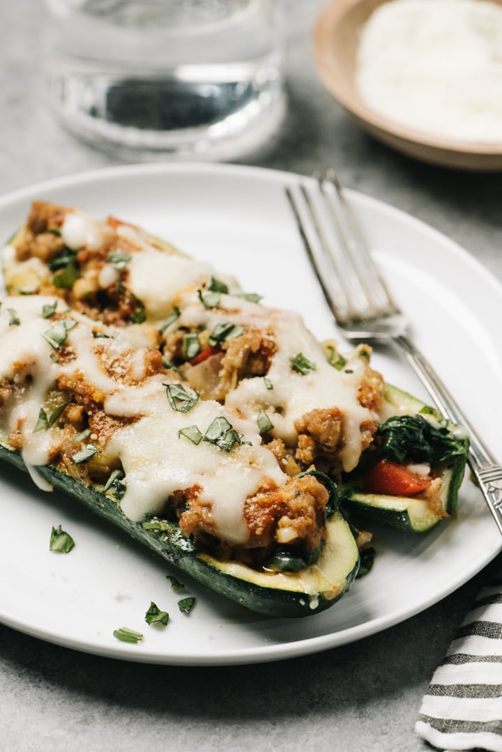 Two italian sausage zucchini boats on a white plate with a silver fork with a glass of water and small dish of parmesan cheese in the background.